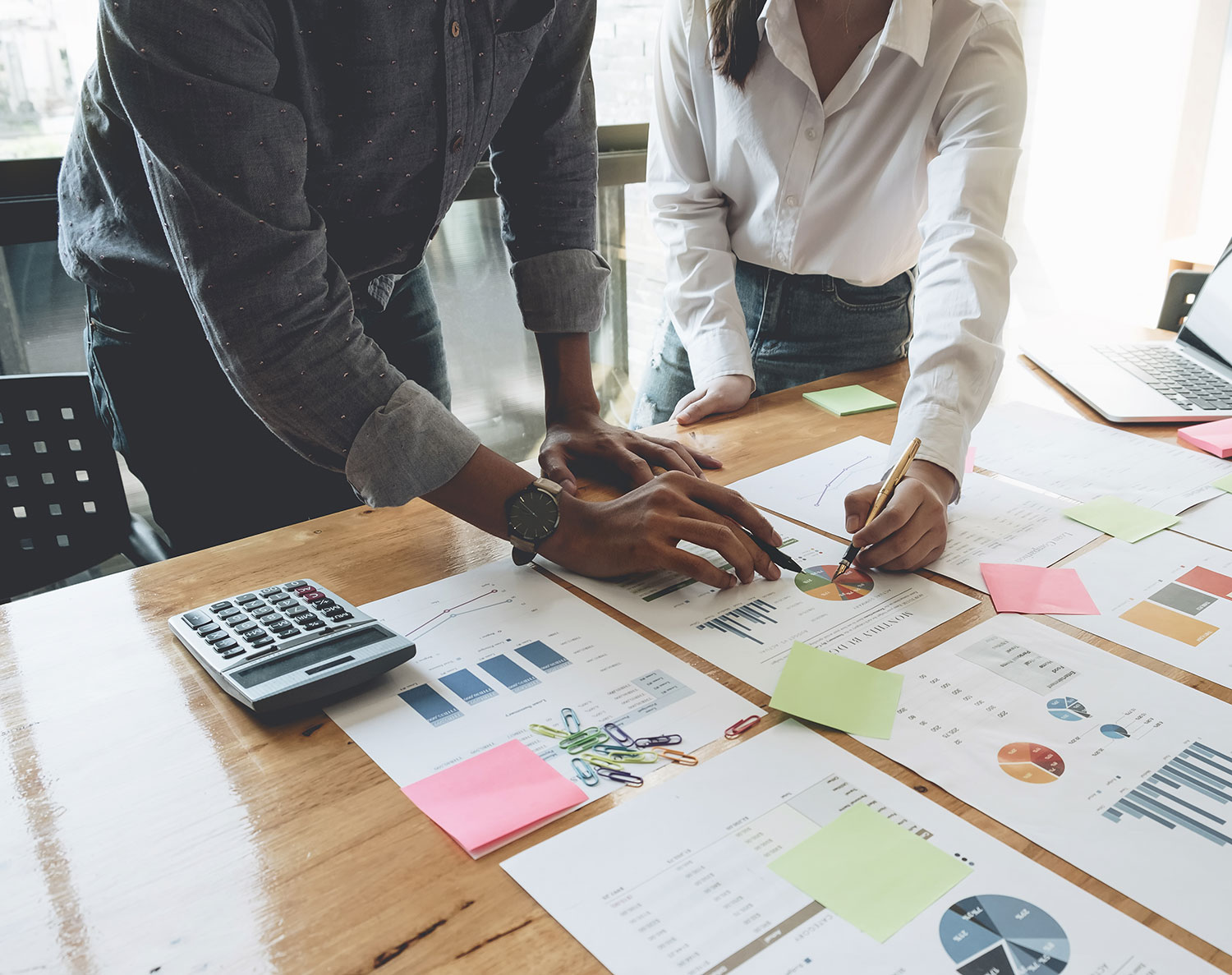 Two business professionals analyzing financial reports with charts and graphs, using sticky notes and a calculator for planning at a collaborative workspace.